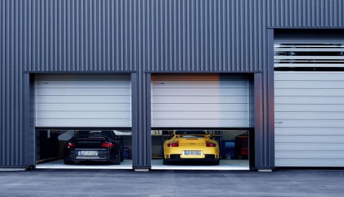Durch die halbgeöffneten Schnelllauftore zweier Garagen sieht man links einen schwarzen und rechts einen gelben Porsche stehen.