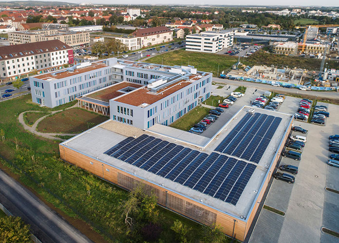 Luftaufnahme des Modulbaus mit Verwaltungsgebäude, großflächiger Photovoltaik-Anlage auf dem Flachdach und angrenzendem Parkplatz. Bild: Kleusberg