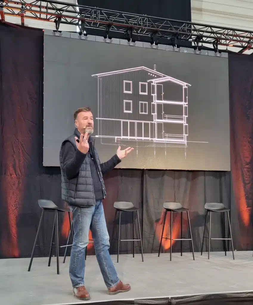 Presenter on stage with a building blueprint displayed behind him on a large screen; man in a dark vest and jeans gesturing while speaking, with stools in the background