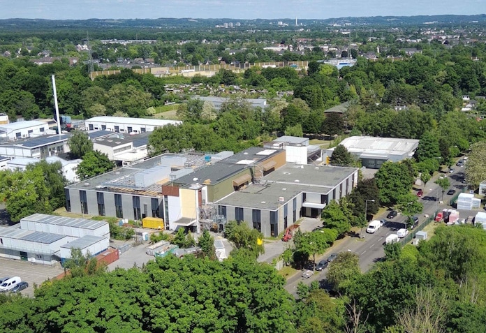 Aerial view of a low-rise industrial complex with flat roofs, parking along a tree-lined street, and surrounding greenery in a town.