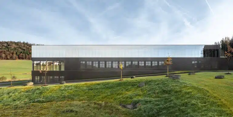 Long modern building with a dark lower level and glass upper facade on a grassy hill under a blue sky.