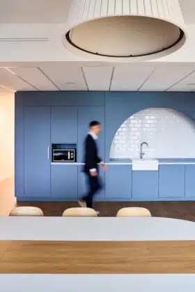 Blurred man in a suit walking past blue kitchen cabinets with built-in microwave and a white sink in a modern showroom style room.