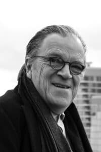Black-and-white portrait of an older man wearing glasses and a scarf, smiling outdoors with buildings in the background.