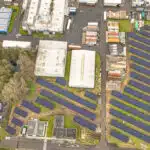 Aerial view of a solar farm with numerous blue panels arranged in rows beside industrial buildings and a river on the right side