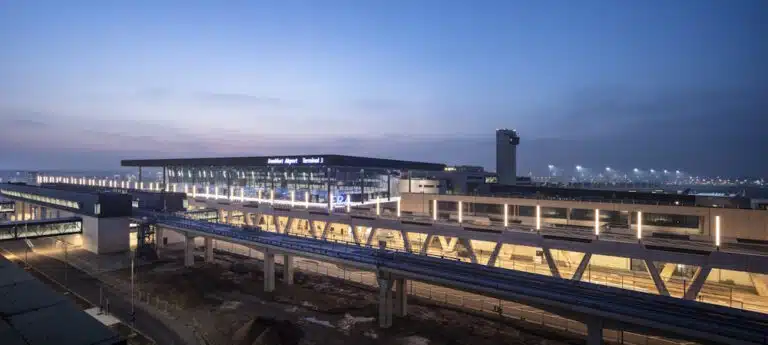 Exterior view of an airport terminal at dusk, with a glass-walled concourse and an illuminated elevated walkway.