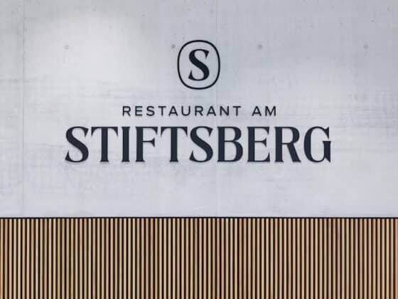 Sign reading 'Restaurant am Stiftsberg' with a circular S logo on a gray concrete wall above a wooden striped panel.