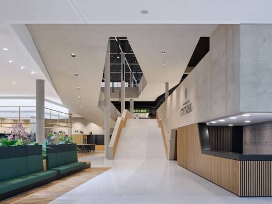 Modern lobby with a central wide staircase, concrete pillars, and green upholstered seating on the left; a wooden reception desk on the right.