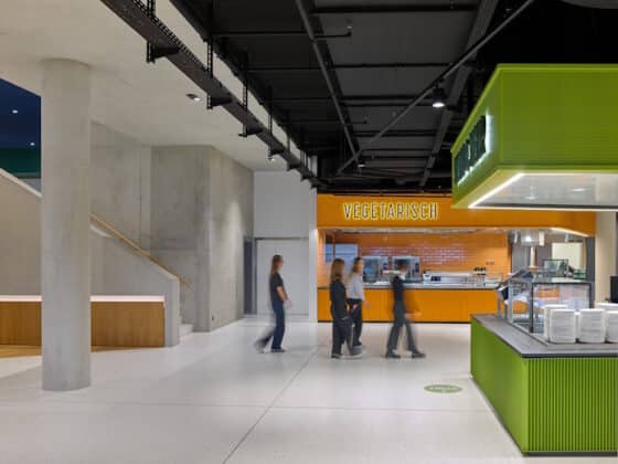Modern cafeteria area with an orange 'Vegetarisch' counter and a green serving station in the foreground; several people walk by in motion blur.
