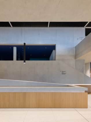 Modern concrete interior with a wooden reception counter, ramp, and a black pendant light in a spacious lobby area.
