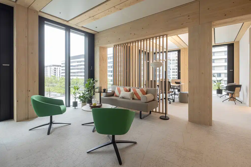 Modern office lounge with green swivel chairs, a beige sofa, wooden columns, and large windows