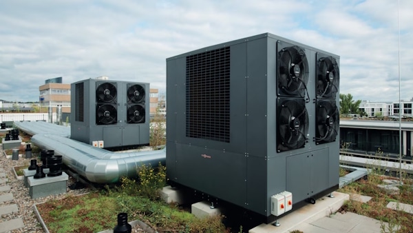 Rooftop view of large industrial cooling units with multiple fans on each side, connected to metal ducts on a flat commercial building roof.