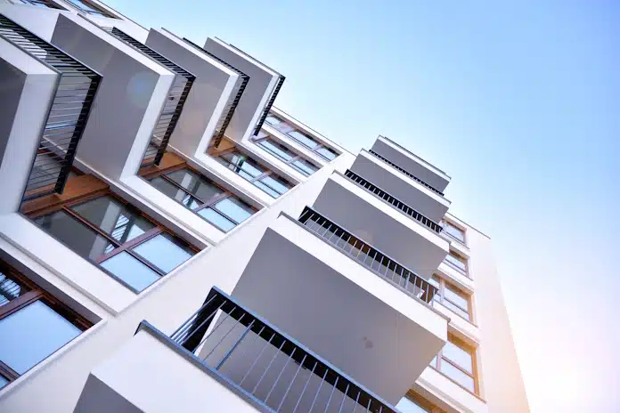 Modern apartment building viewed from below, showcasing balconies and large windows against a clear blue sky.
