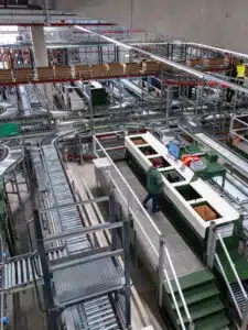 Worker at a green-and-white packing station with a computer, surrounded by a network of conveyor belts in a factory.