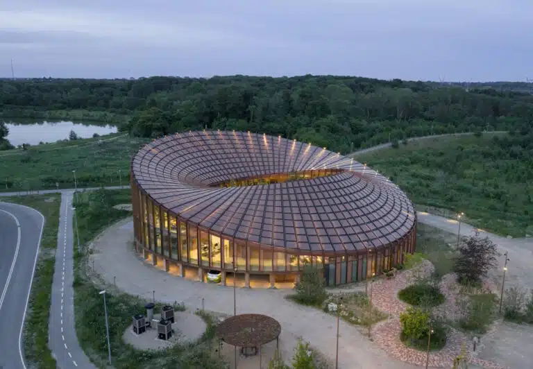 Aerial view of a circular glass-walled building with a grid-pattern roof, surrounded by greenery and a winding road nearby.