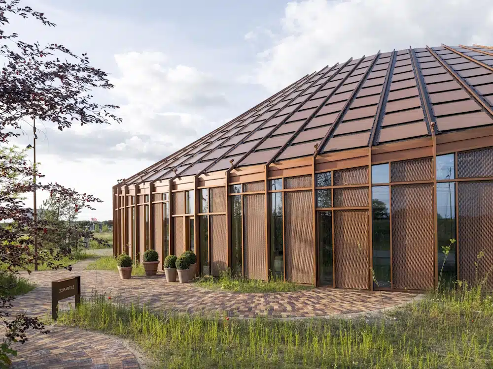 Copper-toned glass pavilion with a curved metal frame, large glass panels, and potted shrubs along a brick walkway outside.