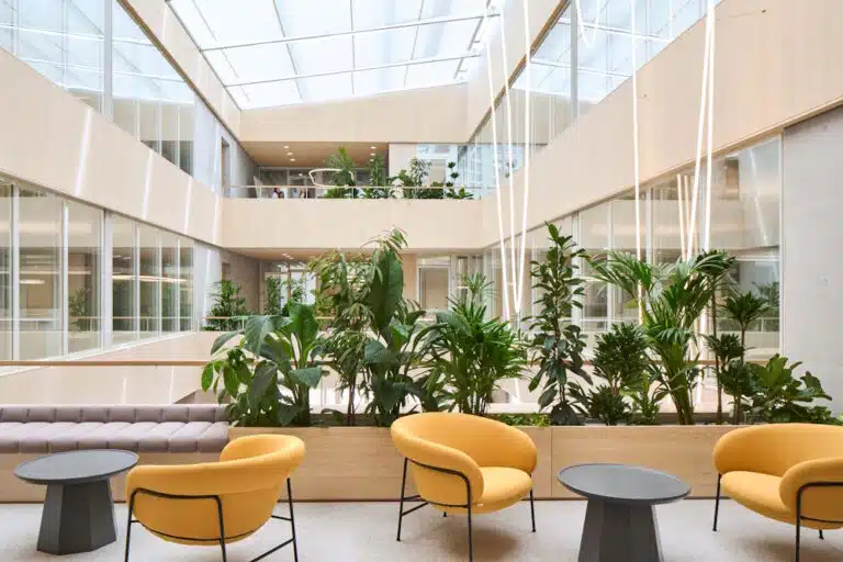 Bright modern atrium with glass ceiling, tall indoor plants, and orange seating around small round tables in an open lounge area. “