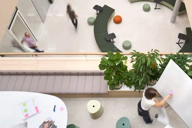 Top-down view of a modern office atrium with curved green seating, plants, and people walking across a light floor area from above.