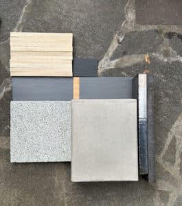 A palette of construction samples on a stone surface: stacked wood slats, dark tiles, gray boards, textured gray square tile, a concrete tile, and a metal ruler.