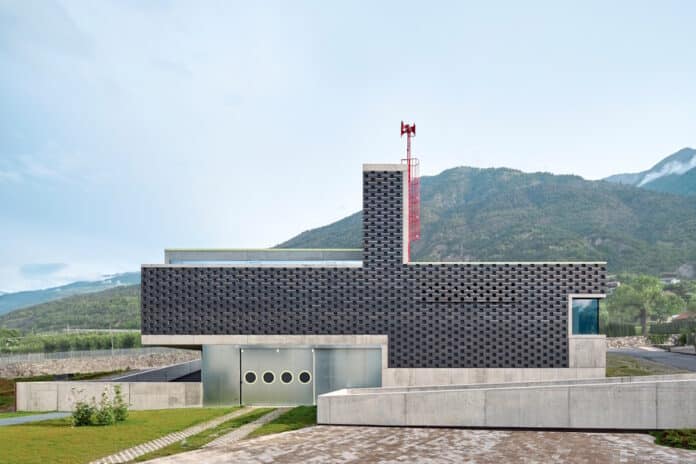 Modern building with a perforated dark brick facade, concrete base, and a red antenna on a hillside with mountains in the background.