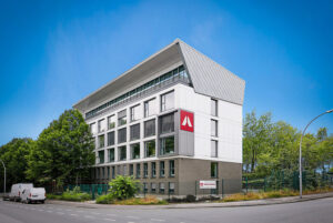 Modern white and gray office building with large windows and a red logo on the corner, by a street with trees under a blue sky