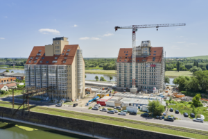 Construction site with two large residential buildings under development beside a river, a tall crane overhead, and a row of parked cars along the road.