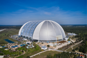 A large white dome building with glass panels on one side, under construction in a green, rural setting (aerial view).