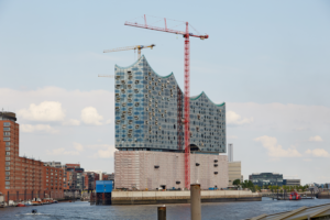 Elbphilharmonie concert hall under construction in Hamburg, with red cranes over the glass-walled, wave-roofed building by the harbor